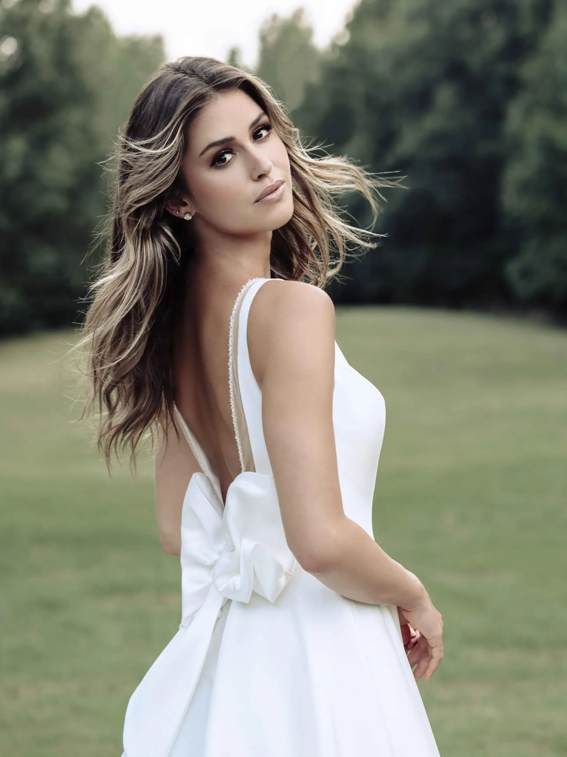 A woman in a white sleeveless dress with a large bow on the back stands outdoors on grass, looking over her shoulder. Her expression is serene.