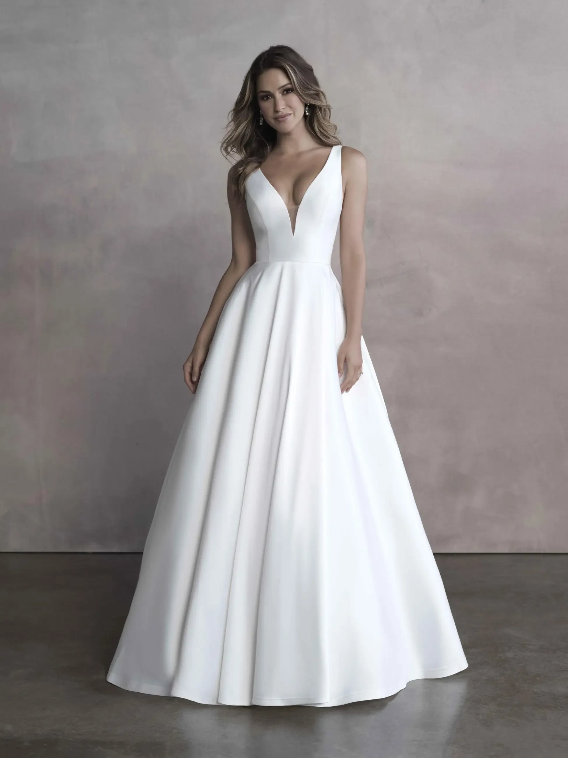 A woman in a simple and elegant white wedding gown stands against a gray background. The dress features a deep V-neck and a full skirt, conveying a timeless and graceful look.