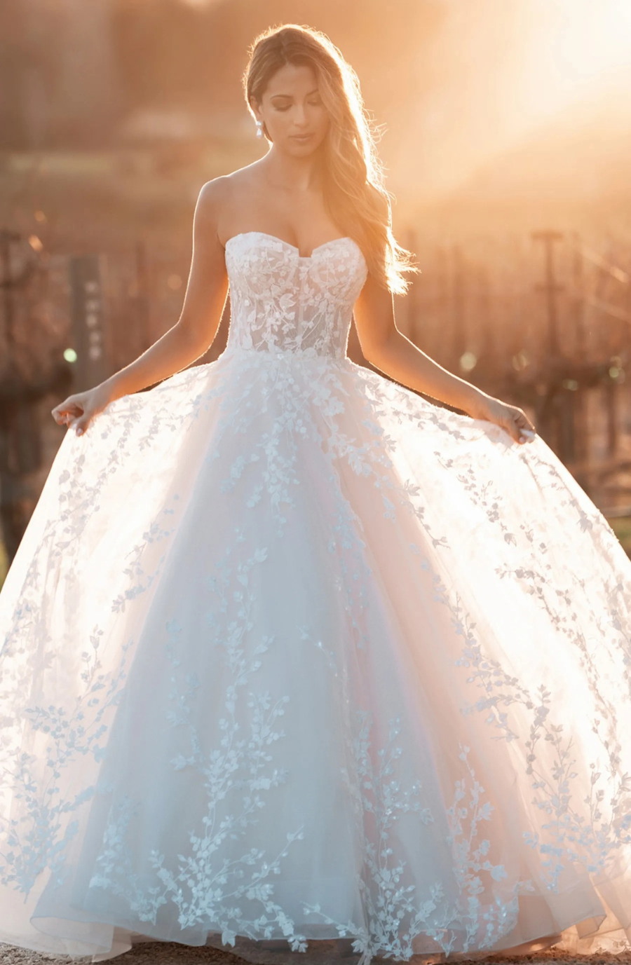 A woman stands outdoors in a flowing white lace wedding gown, illuminated by warm, golden sunlight. Her expression is serene, adding to the dreamy atmosphere.