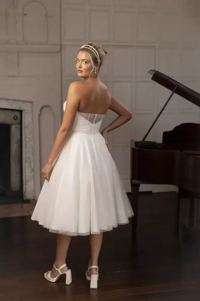 Model is wearing a romantic tea-length tulle gown with a strapless pleated bodice, pearl belt, and softly gathered 50s-inspired skirt.