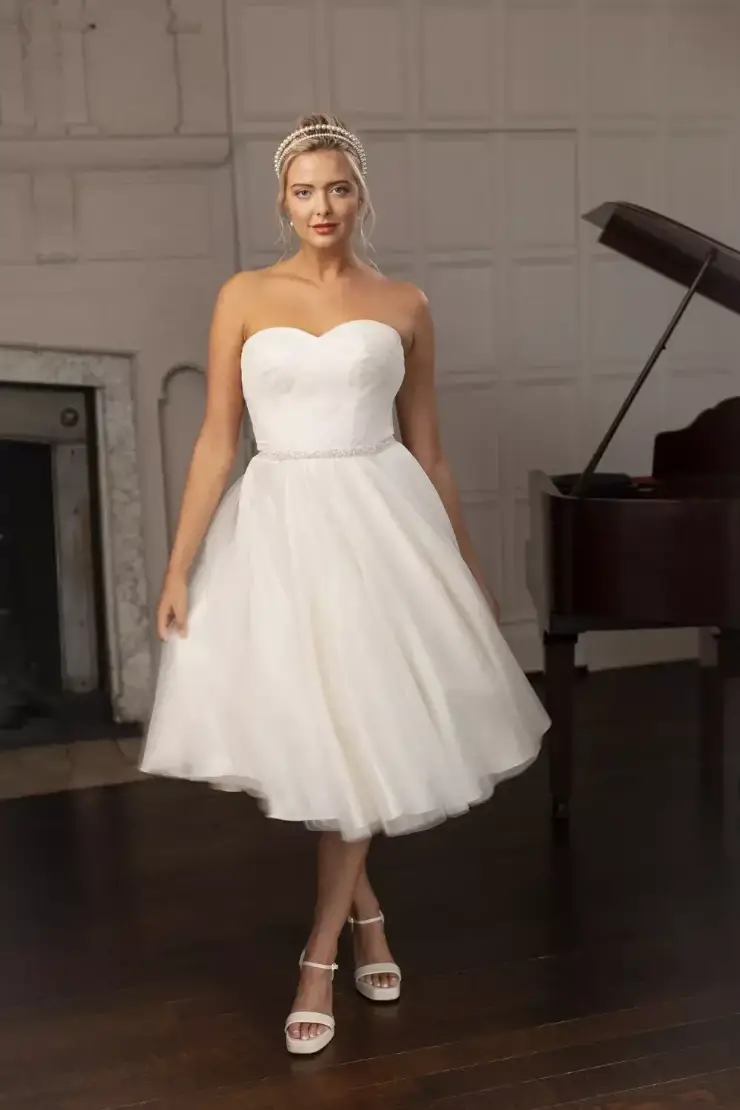 Model is wearing a romantic tea-length tulle gown with a strapless pleated bodice, pearl belt, and softly gathered 50s-inspired skirt.
